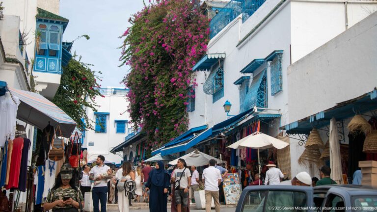 Busy shopping lane beneath cascading magenta bougainvillea, with blue windows, awnings, souvenir stalls, café umbrellas, and pedestrians.