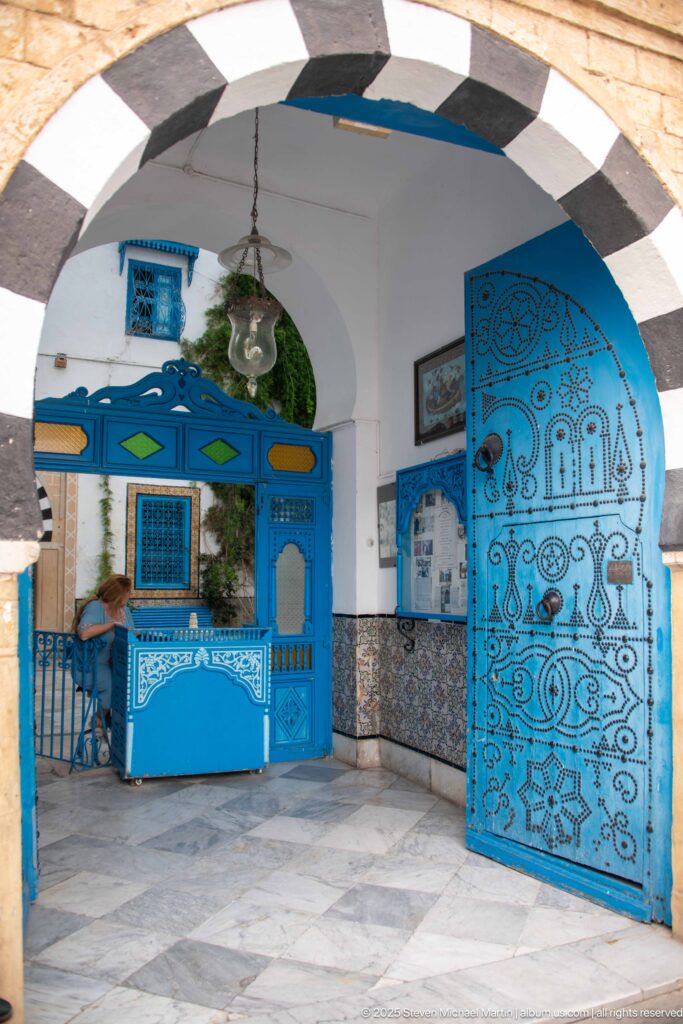 Black-and-white striped arch opening onto blue wrought-metal doors, tiled walls, hanging lantern, and greenery in a Sidi Bou Said courtyard.