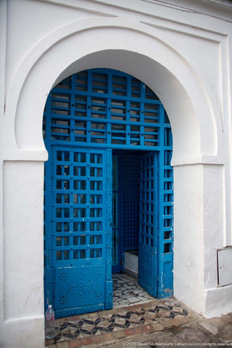 White horseshoe arch framing open blue lattice doors above a patterned threshold and shaded vestibule.
