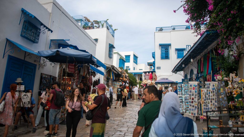 Crowded cobbled lane lined with souvenir stalls, white buildings, blue trim, awnings, and bougainvillea.