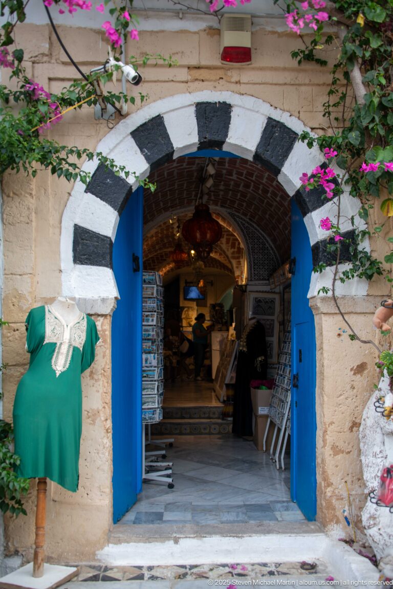 Striped masonry arch with open blue doors leading into a small shop filled with merchandise and hanging lamps.