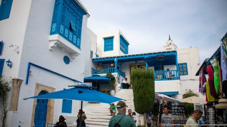 White buildings with blue balconies, shutters, railings, and awnings around stone steps crowded with pedestrians.