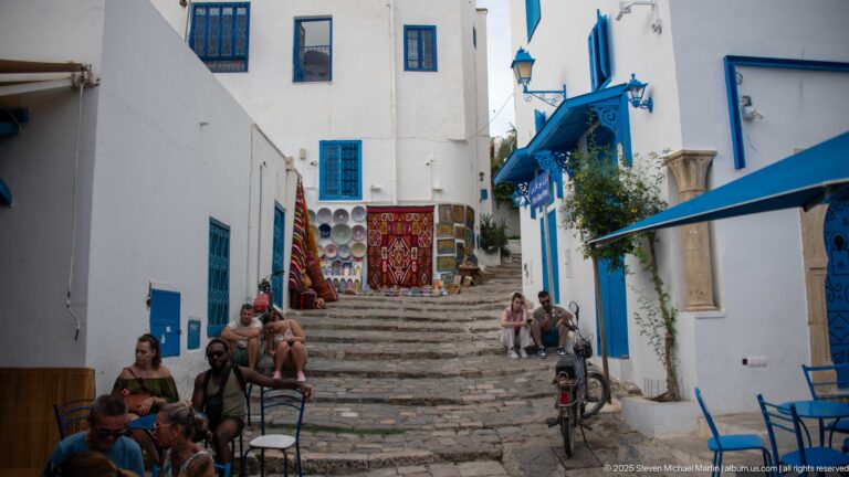 Outdoor café tables beside a steep stone stair lined with white buildings, blue trim, shops, and pedestrians.