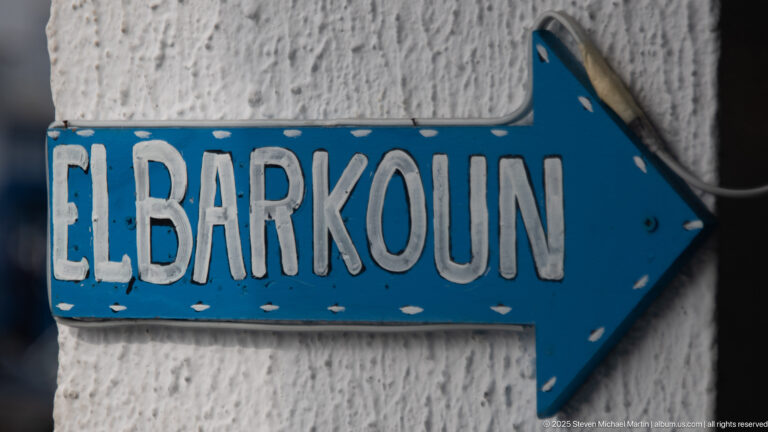 Blue arrow-shaped sign reading “EL BARKOUN” in white letters, mounted on a rough white wall and pointing right.