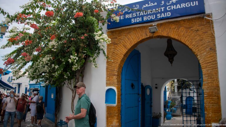 Arched blue entrance to Café Restaurant Chargui beneath a bilingual sign, with flowering branches and visitors outside.