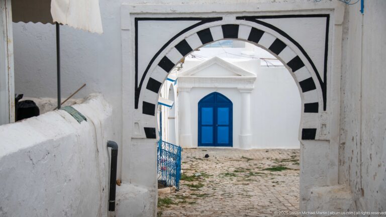 Black-and-white striped arch framing a white façade with a bright blue double door and cobbled ground.