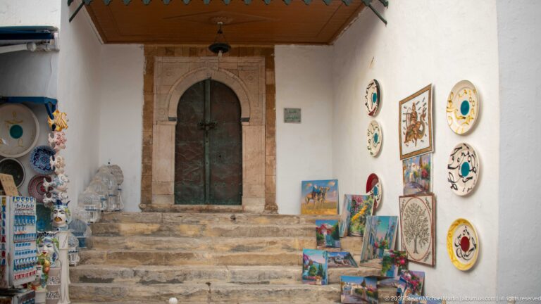 Painted ceramic plates and small artworks displayed on stone steps beside a tall arched wooden doorway.