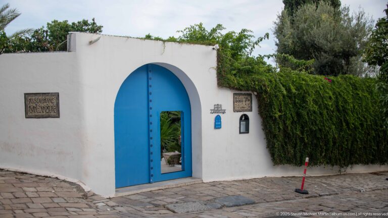 Rounded white garden entrance with a tall blue gate, small plaques, lantern, and dense greenery beyond.