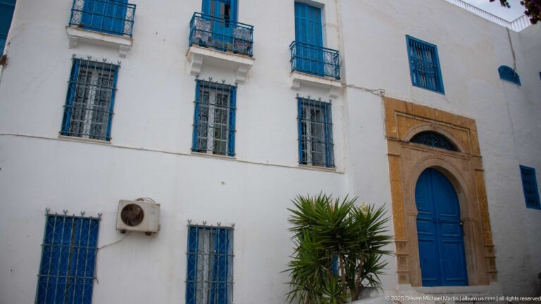 White multi-story façade with blue barred windows, small balconies, and a carved stone portal around a blue door.