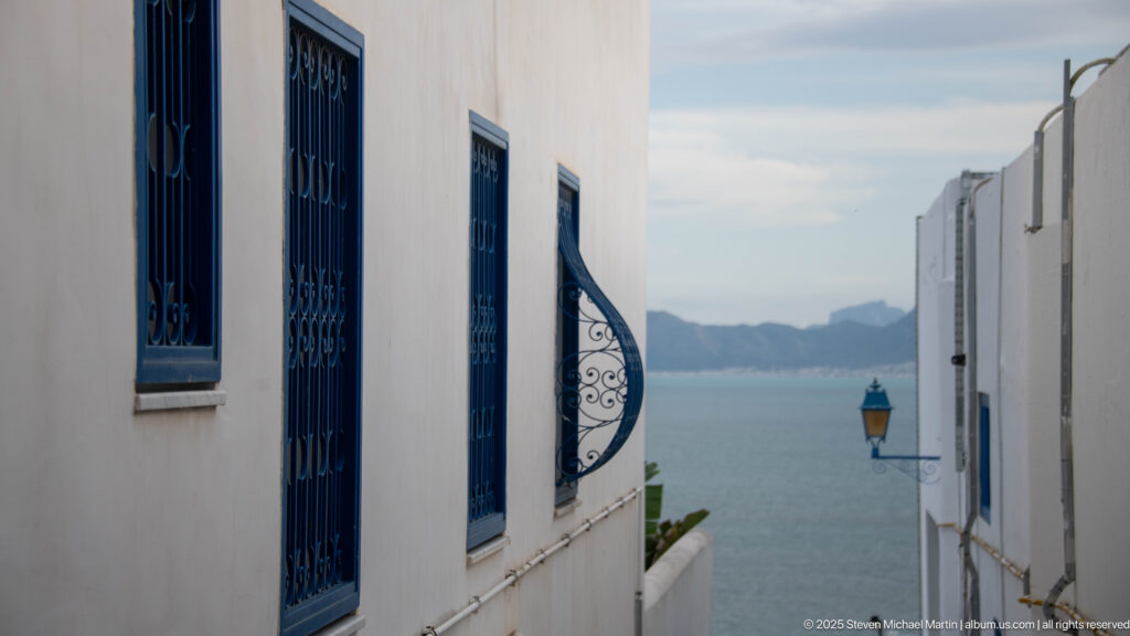 Calm sea and distant coast framed by narrow white walls with blue window grilles and a wall lantern.