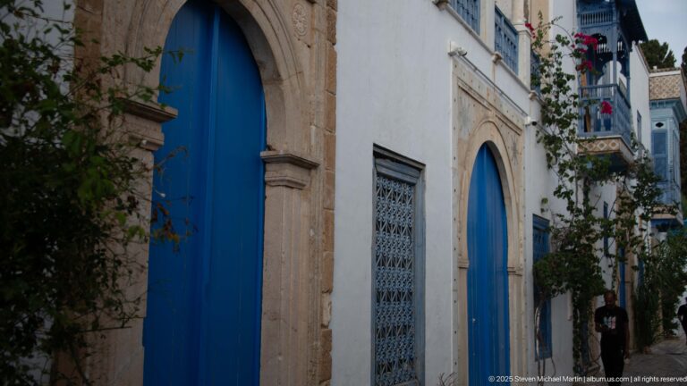 Two blue arched doors and a large metal-grilled window set in a white façade with stone surrounds.