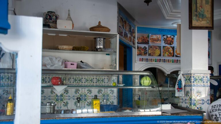 Small food counter with patterned tiles, glass display case, open shelves, blue trim, and a wall menu of dish photographs.