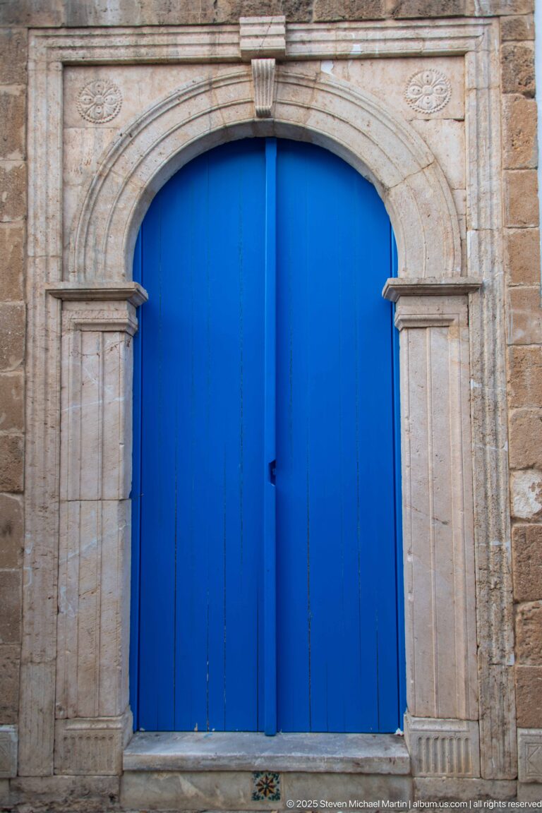 Large blue double doors beneath a shallow carved stone arch with pilaster-like side elements and weathered paint.