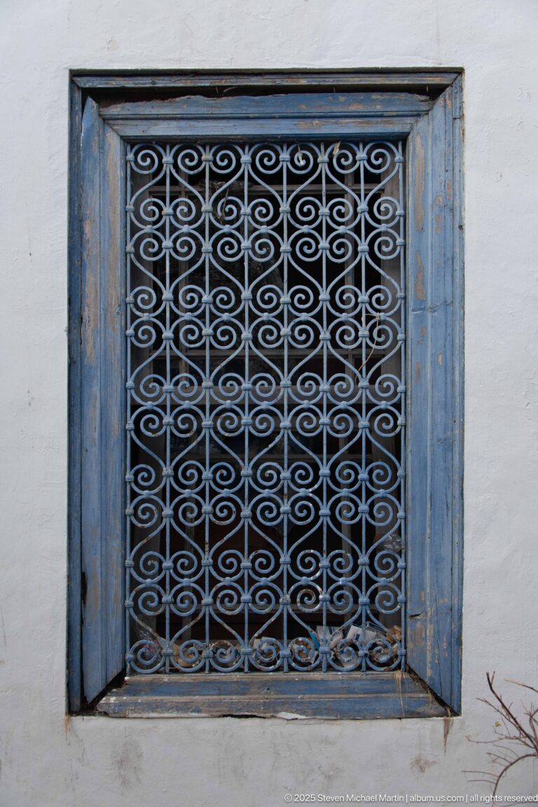 Blue wooden window frame with an ornate repeating iron scroll grille set in a white plaster wall.