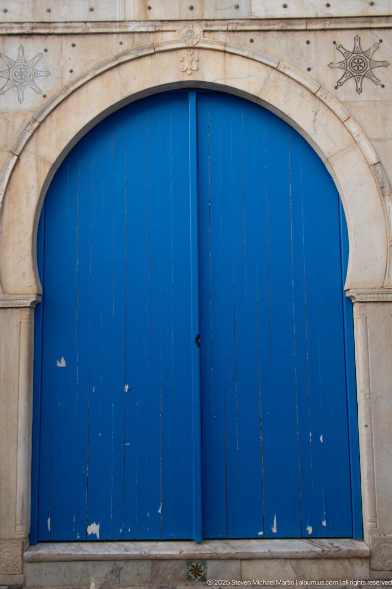 Wide blue plank double doors set beneath a pale stone arch with incised ornament and visible weathering.