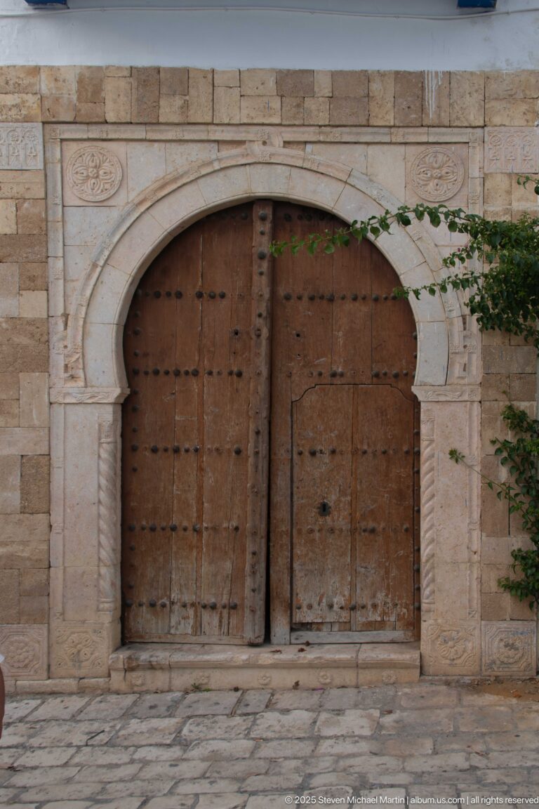 Weathered wooden double doors with iron studs framed by a carved pale-stone arched portal.