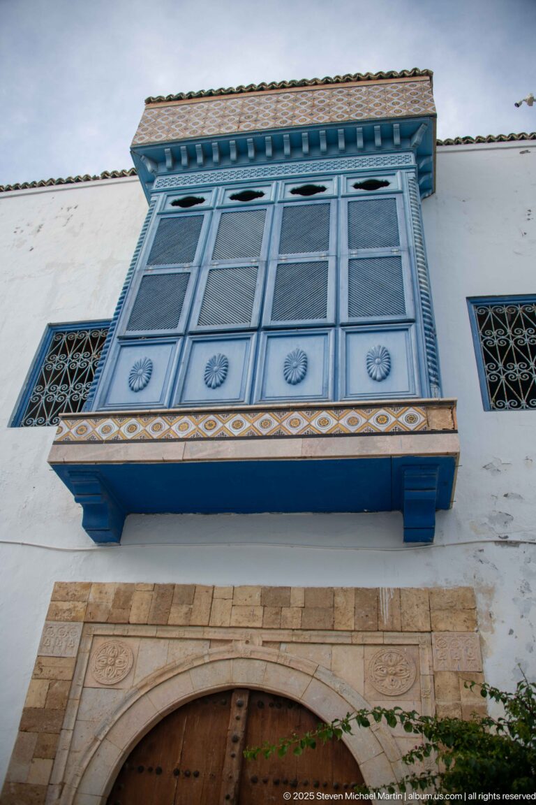Blue enclosed projecting balcony with screened panels and decorative brackets above a carved stone doorway.