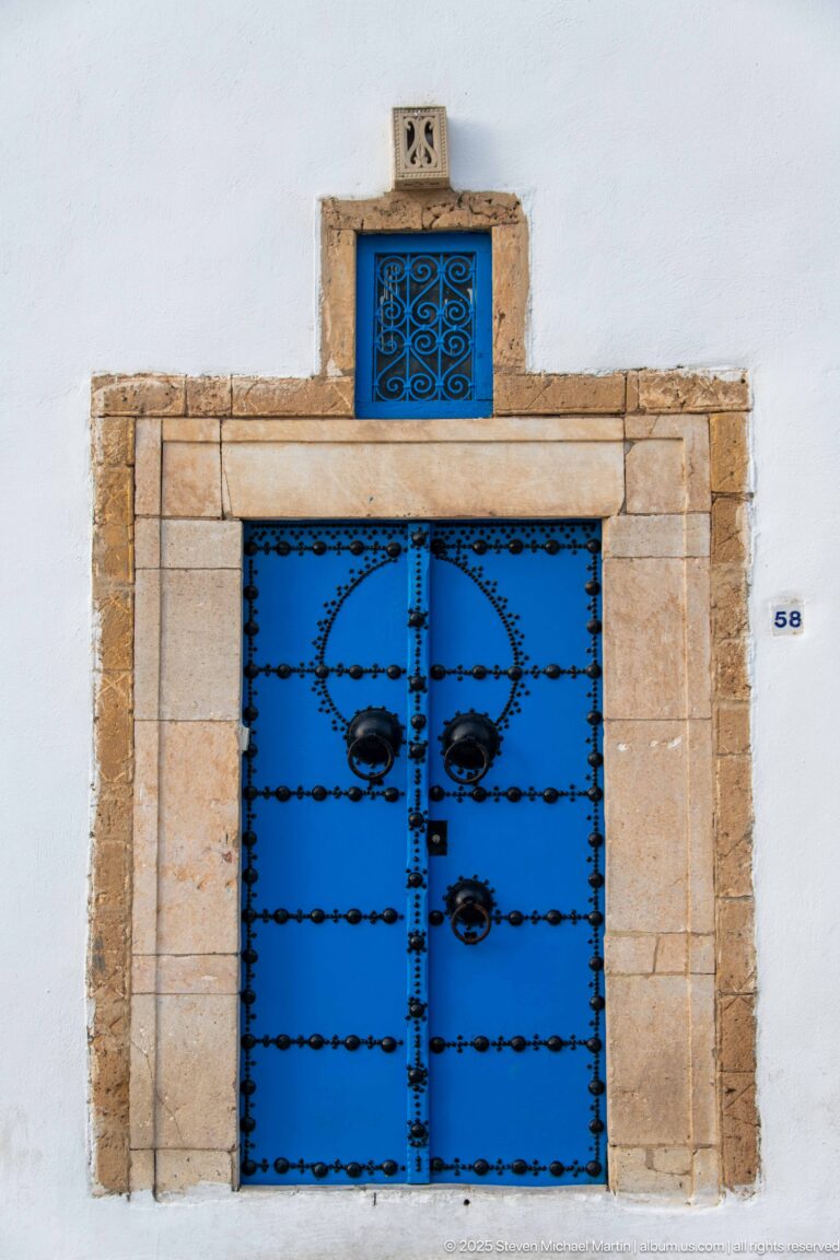 Blue studded double doors with circular iron bosses and a small grilled transom in a pale stone frame.