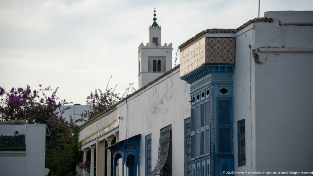 Blue projecting balcony on a white street façade with a slender white mosque minaret rising behind it.