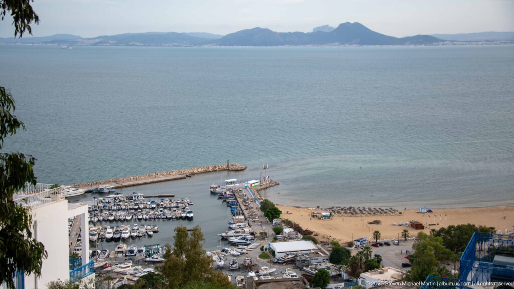 High view over Sidi Bou Said marina, breakwaters, sandy beach, calm bay, and distant mountain coastline.