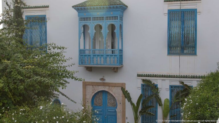 White façade with an ornate blue projecting window above a blue arched door, surrounded by barred windows and foliage.