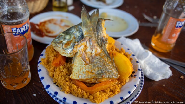 Whole cooked fish standing on couscous with vegetables and a lemon wedge, surrounded by plates and orange soft-drink bottles.
