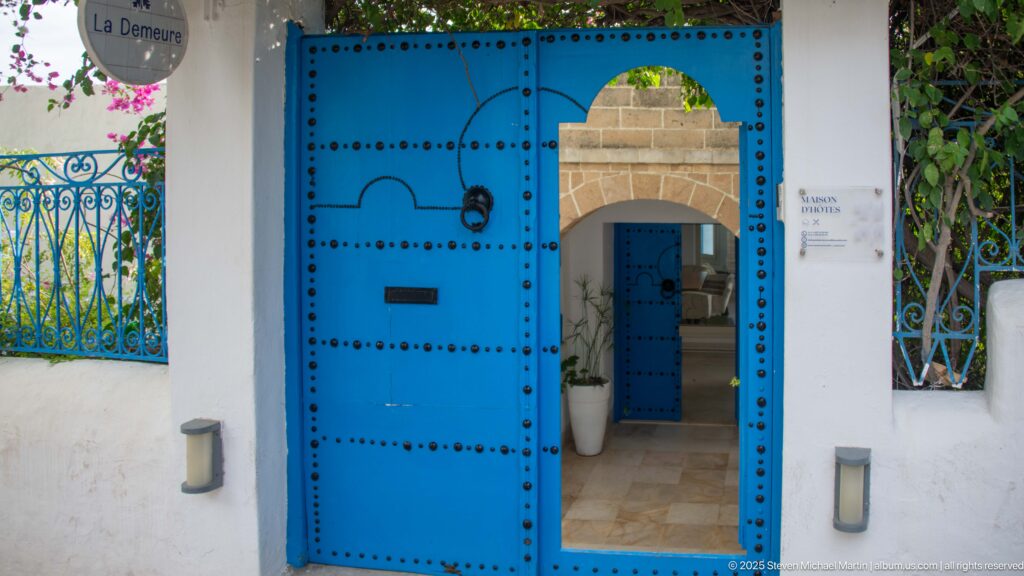 Large studded blue gate partly open to a tiled courtyard and smaller blue doorway, beside a blue iron fence.