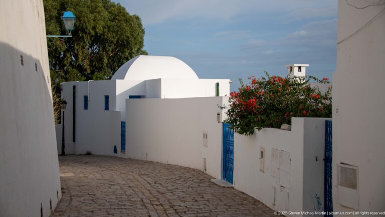 Cobbled lane curving beside a white domed house with blue gates, small wall openings, and flowering bougainvillea.
