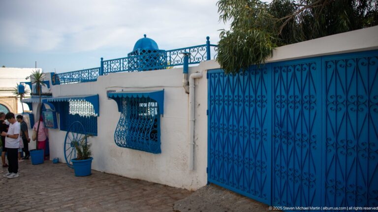 White façade with ornate blue iron gates, window grilles, railings, potted plants, and a small blue rooftop dome.