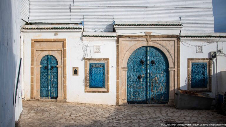 White façade with central arched blue double doors, side doorway, and barred blue windows in pale stone frames.