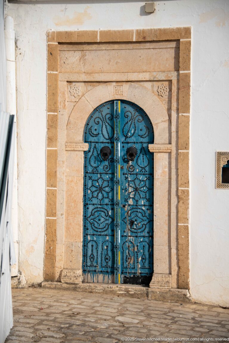 Blue wrought-iron double doors with scrollwork and circular bosses set in a pale carved stone arch.