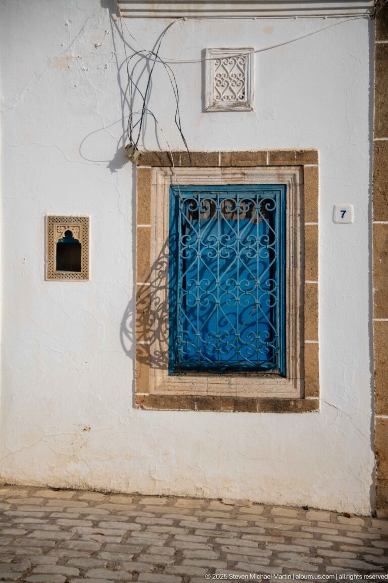 Blue decorative metal window grille in a pale stone frame on a white wall, with overhead wires and small wall niche.