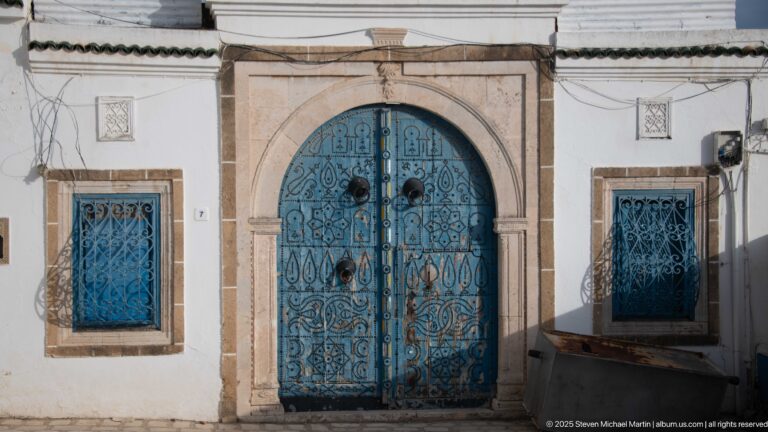 Tall arched blue double doors with ornate ironwork and circular bosses, flanked by blue barred windows in stone frames.