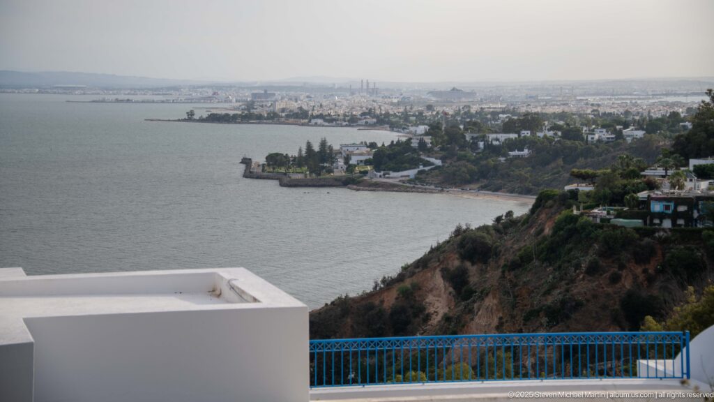 Elevated coastal view over a broad bay, wooded promontory, urban shoreline, and distant industrial skyline from a blue-railed terrace.