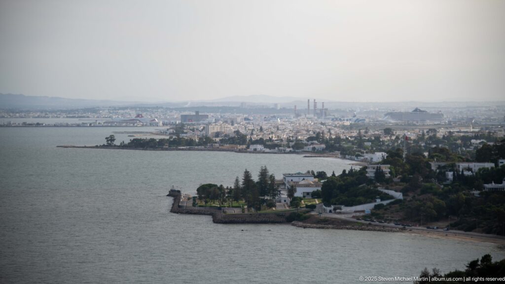 Hazy panoramic view across the bay toward dense urban development, industrial stacks, and a low waterfront peninsula.