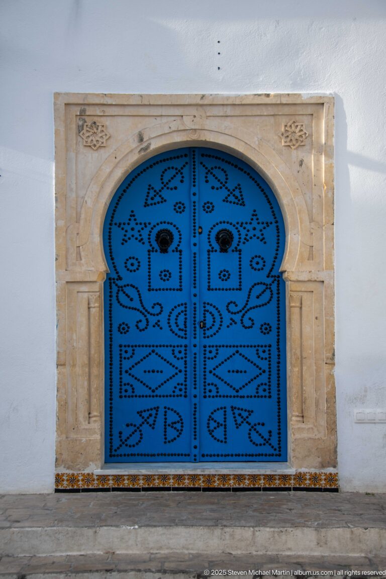 Blue arched double doors covered with geometric stud patterns, framed by pale stone and a narrow tiled base.