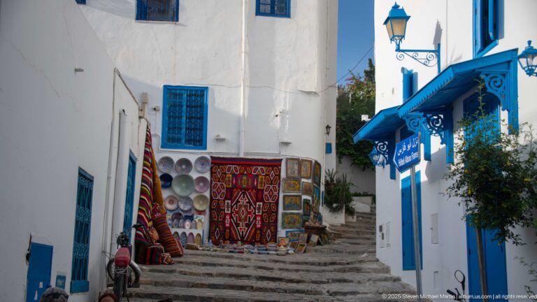 Stone steps between white-and-blue buildings leading to a souvenir display of rugs, ceramics, and other goods.