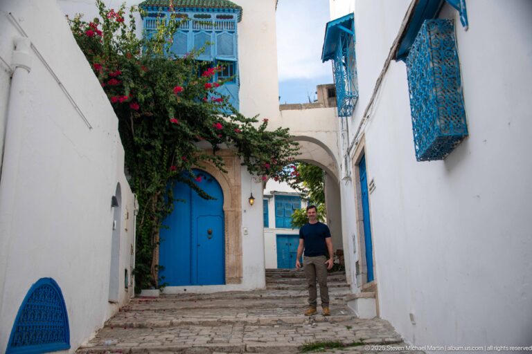 Man standing on stone steps between white walls with blue doors, projecting blue grilles, and bougainvillea.