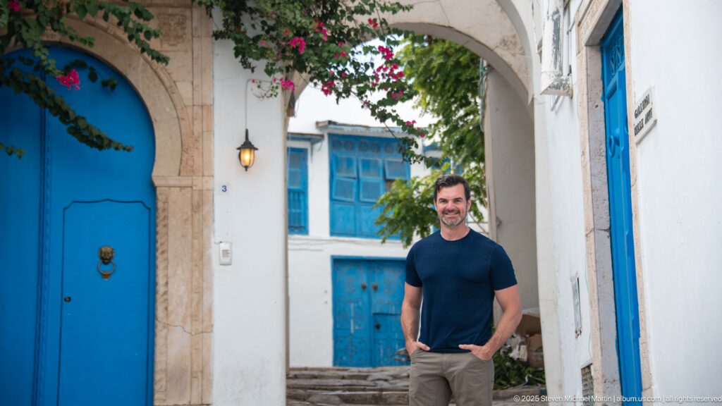 Man standing beneath a pale stone arch beside a large blue door, with blue windows and steps behind him.
