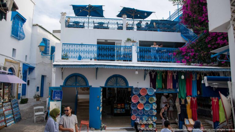 Small commercial courtyard with blue balconies, arched shopfronts, rooftop umbrellas, souvenir displays, visitors, and bougainvillea.