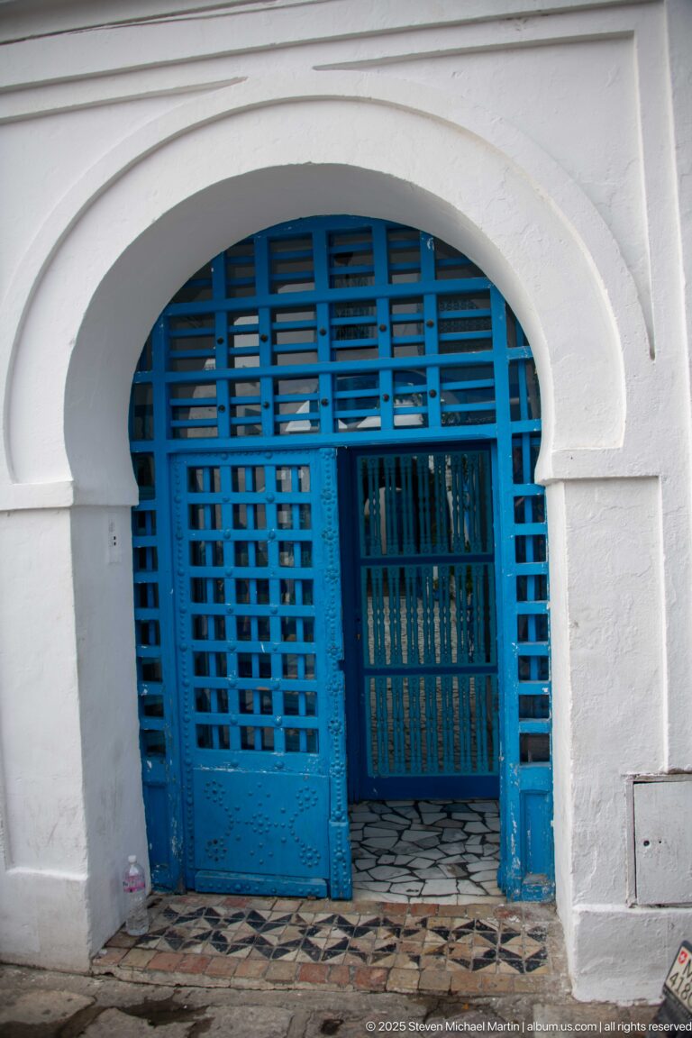 Broad white arch filled with blue wooden lattice panels and a darker inner grille above patterned floor tiles.