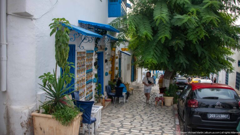 Tree-shaded cobbled lane with white-and-blue shops, small café tables, pedestrians, and a parked car.
