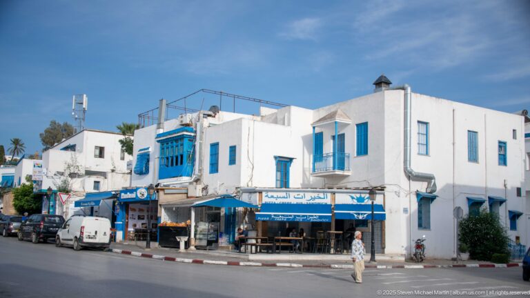 Street corner with white buildings, blue shutters and awnings, café seating, storefronts, parked vehicles, and a pedestrian crossing.
