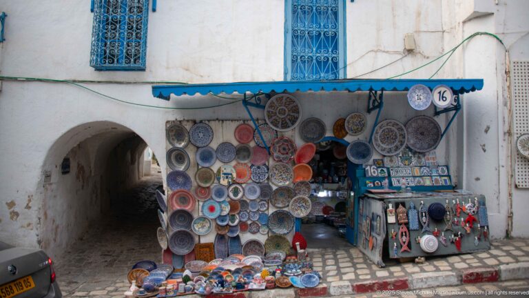 Shopfront densely covered with painted ceramic plates beside a low arched passage, blue awning, and goods on the pavement.