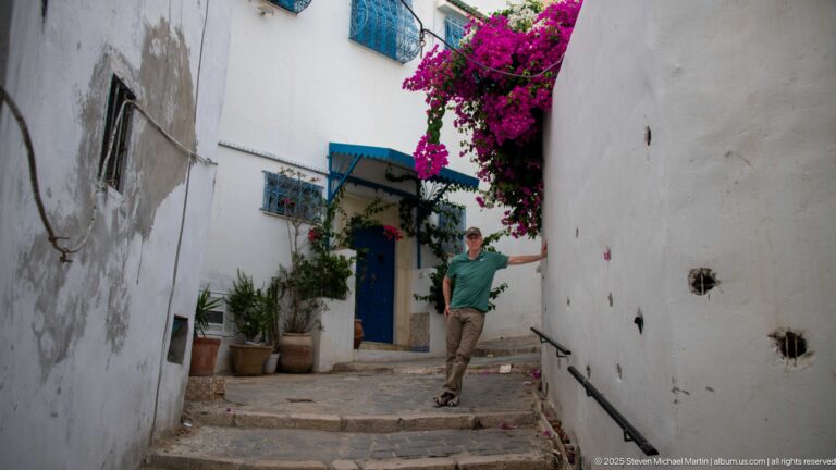 Man standing on narrow stone steps between white walls, with a blue doorway and bougainvillea above.