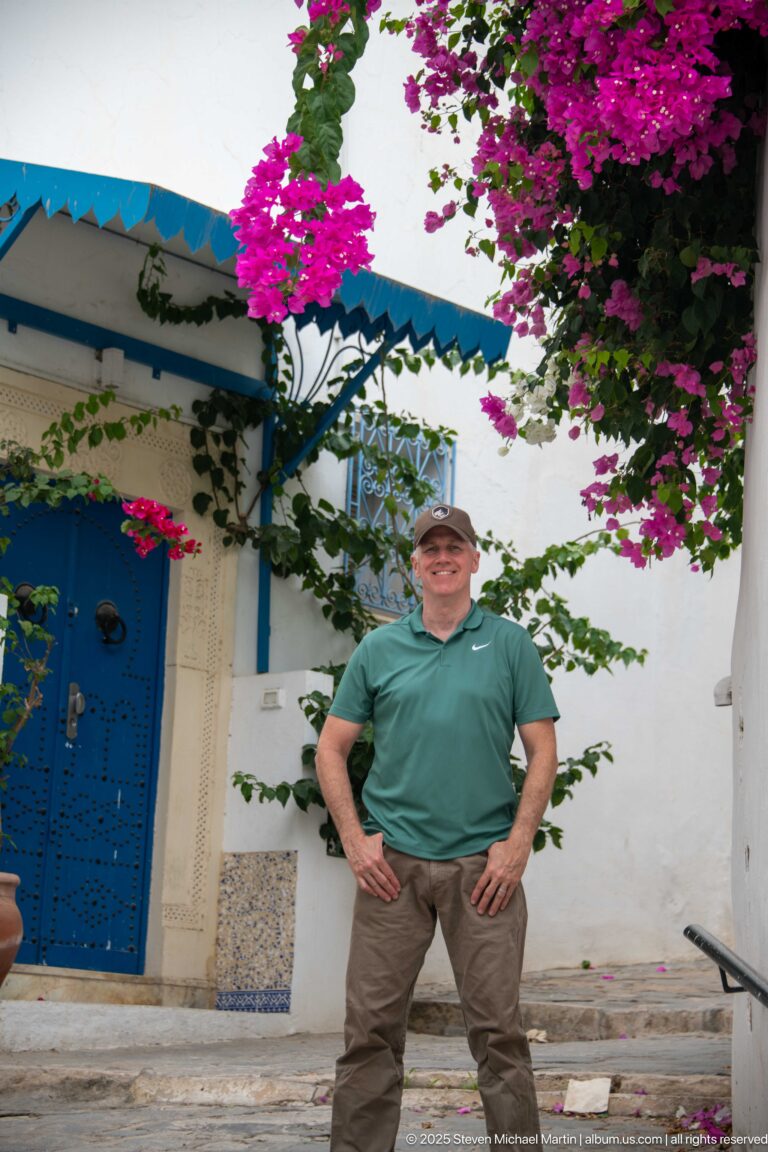 Man standing beside a patterned blue arched door under dense magenta bougainvillea on a white façade.