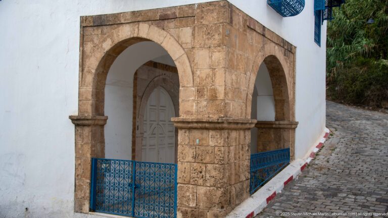 Warm stone corner arcade with two rounded arches, recessed white door, blue railing, and cobbled street curving uphill.