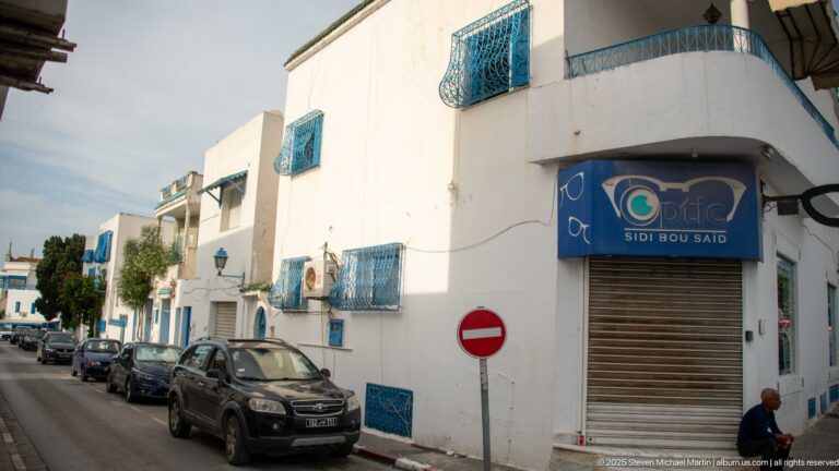 Closed Optica Sidi Bou Said storefront on a white corner building with blue grilles, parked cars, and a red no-entry sign.