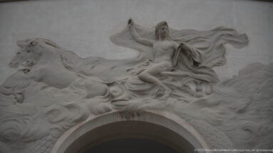 White sculptural relief of a draped female figure with horses and billowing fabric above an arch on the Municipal Theatre.