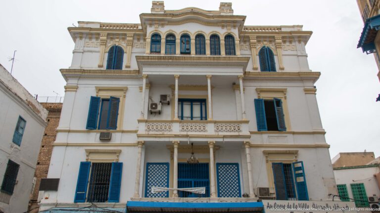 White Tunis building with blue shutters, columned balconies, arched upper windows, and decorative parapet.